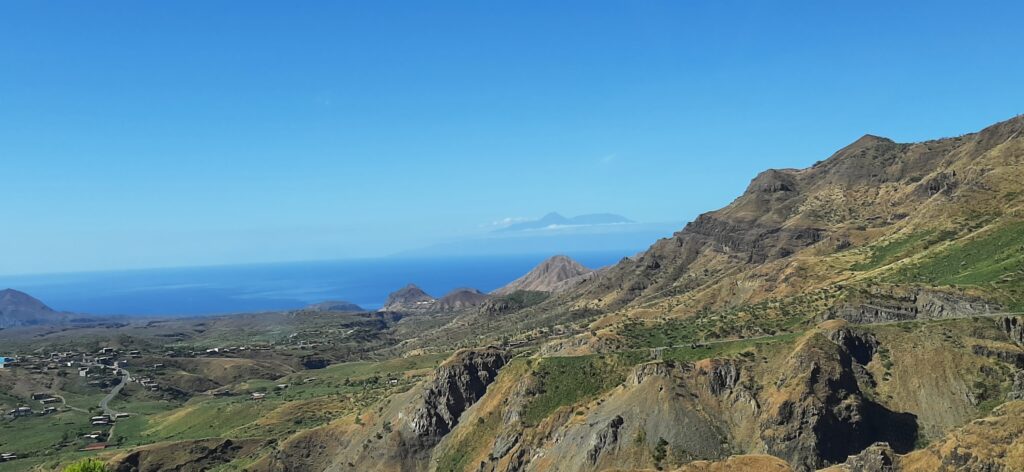 A panoramic view of a rugged, arid landscape with deep valleys and sharp ridges. The Atlantic Ocean is visible in the distance, and the outlines of two islands can be seen on the horizon under a clear blue sky. A small village is nestled in the valley on the left.