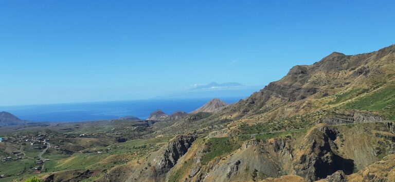 A panoramic view of a rugged, arid landscape with deep valleys and sharp ridges. The Atlantic Ocean is visible in the distance, and the outlines of two islands can be seen on the horizon under a clear blue sky. A small village is nestled in the valley on the left.