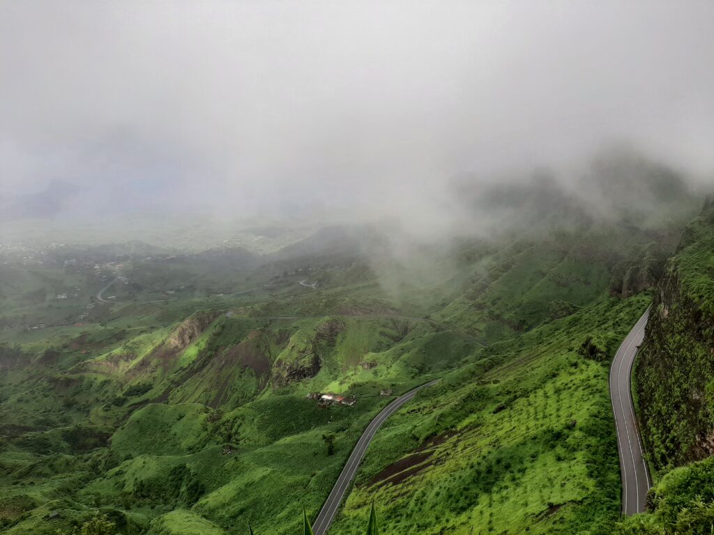 A winding road cuts through a steep, verdant landscape shrouded in thick mist. The hillsides are a vibrant green, with patches of terraced fields. The road snakes down into a foggy valley where a small house is visible
