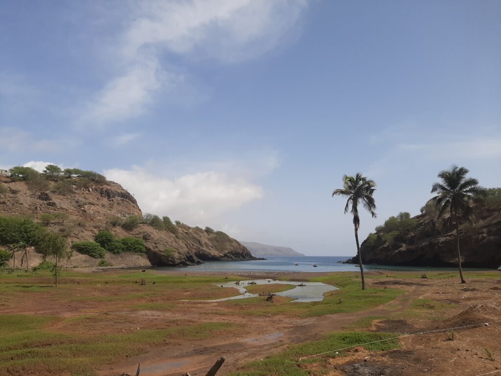 view of a coastal inlet with a small stream winding through a grassy, brown field toward the sea. Two palm trees stand on the banks near the water. The inlet is flanked by rocky cliffs, and a small boat is anchored in the calm water