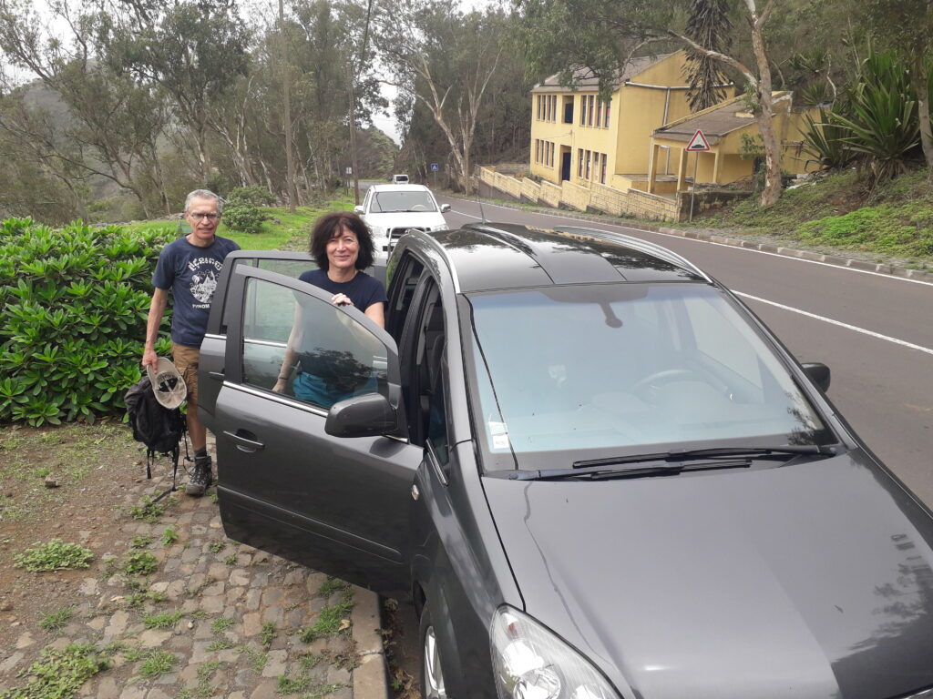 A man and a woman stand next to a gray minivan with its front door open on a cobblestone road. A yellow building is in the background, surrounded by lush green foliage.