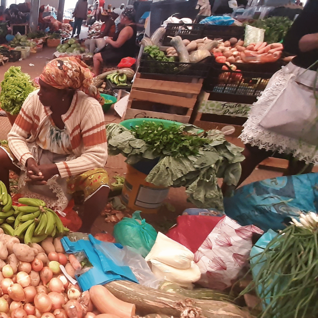 Bustling indoor market in Assomada, Cape Verde — vendor seated among bananas, onions, leafy greens, carrots and other fresh produce, with shoppers in the background